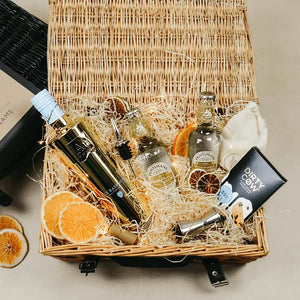 A personalised gift hamper containing a gold bottle of AU Vodka with Blue Raspberry, premium Indian tonic water, fairy lights, and a bar of chocolate, all presented in a wicker basket with a black label and orange fruit.