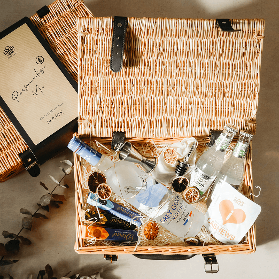 A wicker  corporate gift hamper basket open, displaying an assortment of premium spirits, tonics, snacks, garnishes, and barware, with a personalized message sleeve