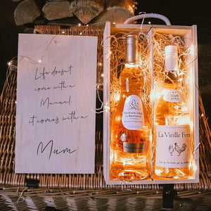 Two bottles of wine in a decorative box with a card reading 'Life doesn't come with a manual, it comes with a mum' on a wooden surface.