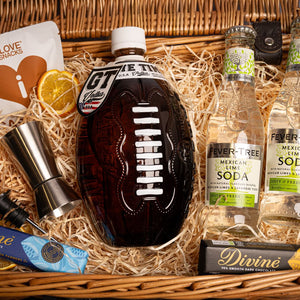 Close up of a Vodka bottle shaped like an NFL ball within a gift hamper with wood wool soda water mixer bottles, barware and some snacks